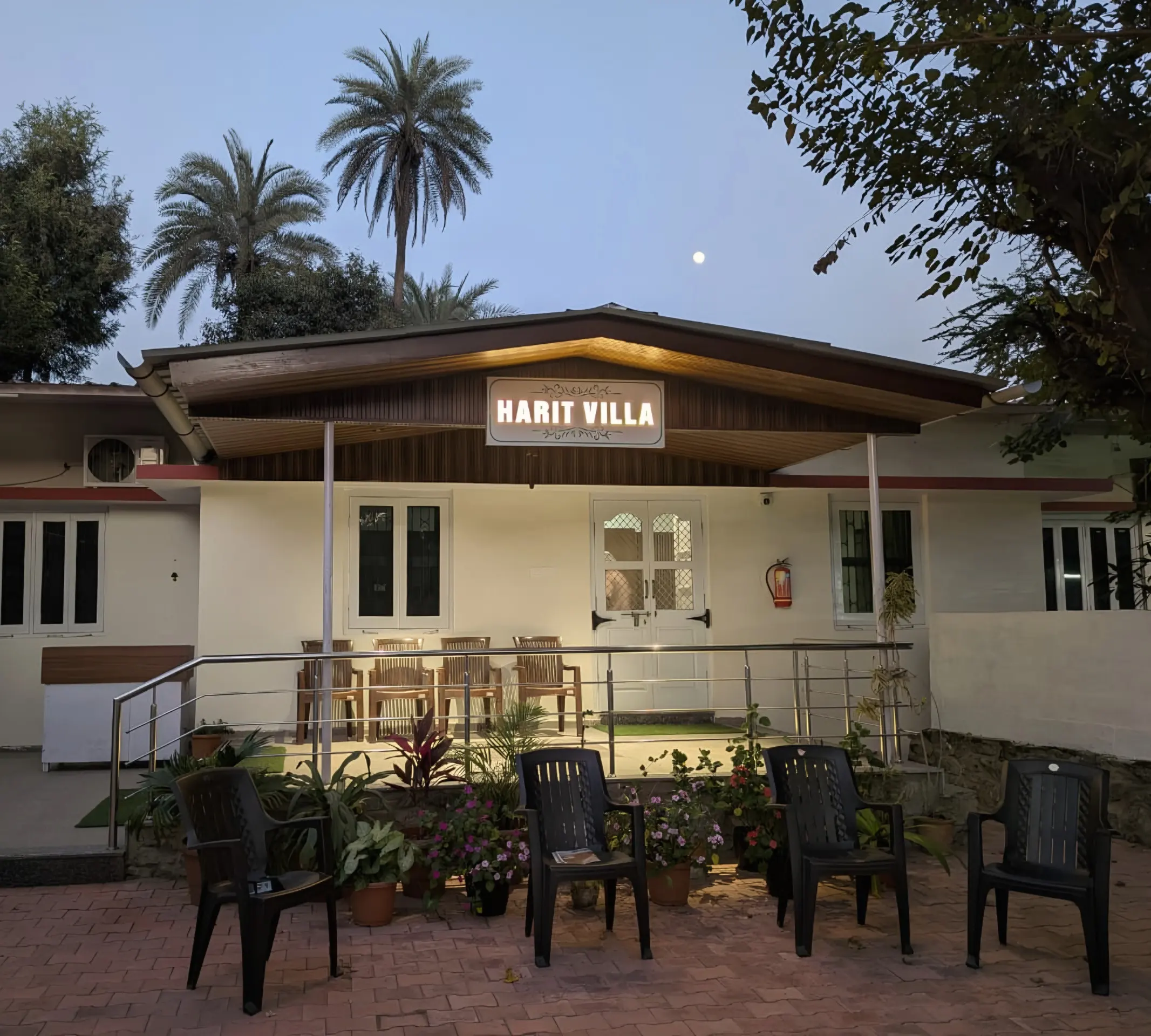 Harit Villa at dusk, with its lit sign, palms, a full moon and flowers along the courtyard